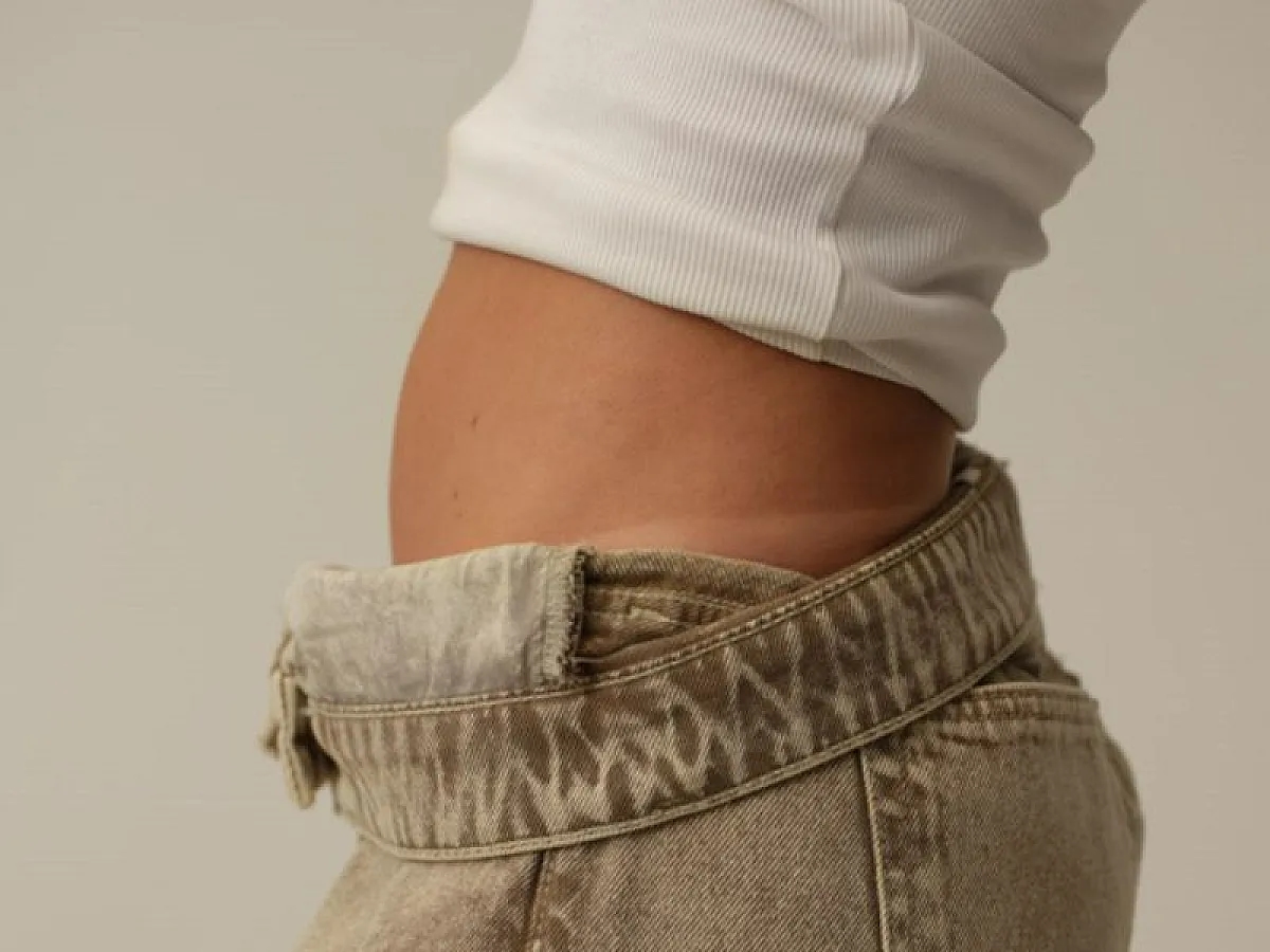 Frau trägt weisses Unterhemd zu grauer Jeans und zeigt Tan Lines am Becken zum Thema Tanrevel-Selbstbräuner